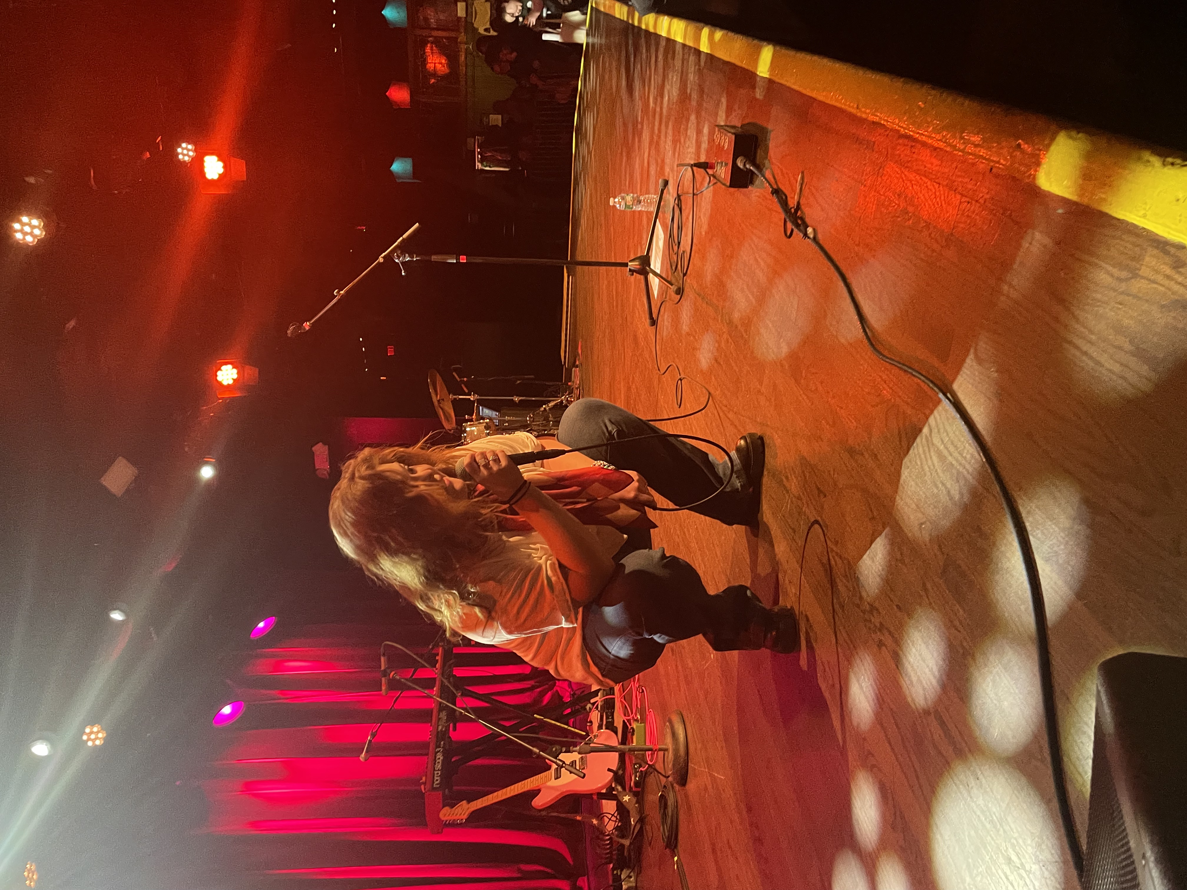 young woman crouching on a concert stage, singing into a microphone. Orange and red lights surround her